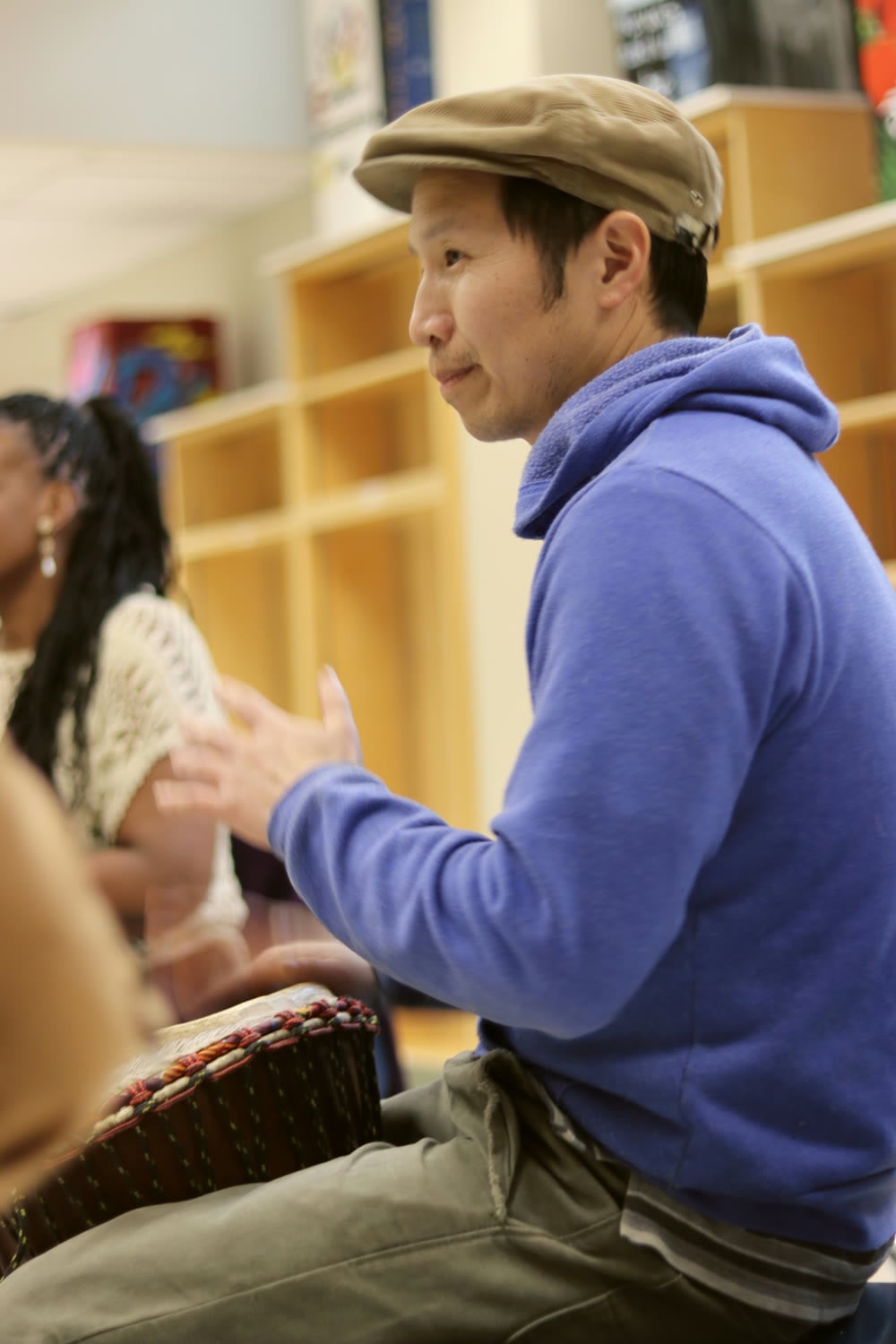 Marcus Fung teaching hand drumming, smiling, in a flat cap